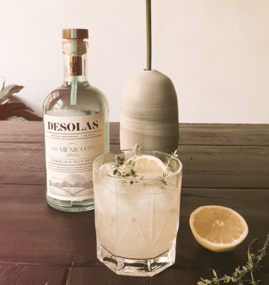 A rocks cocktail in a short glass with a bottle of Desolas Mezcal behind it