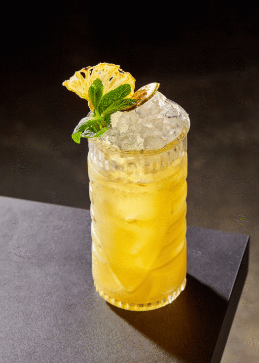 A tall glass with a yellow cocktail over fresh ice and a black background.