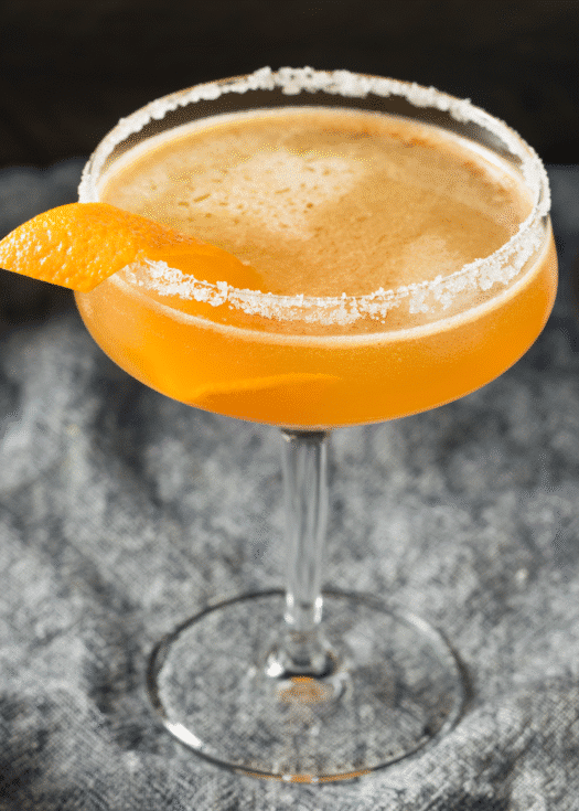 An orange colored cocktail in a coupe glass with a salted rim and an orange peel garnish
